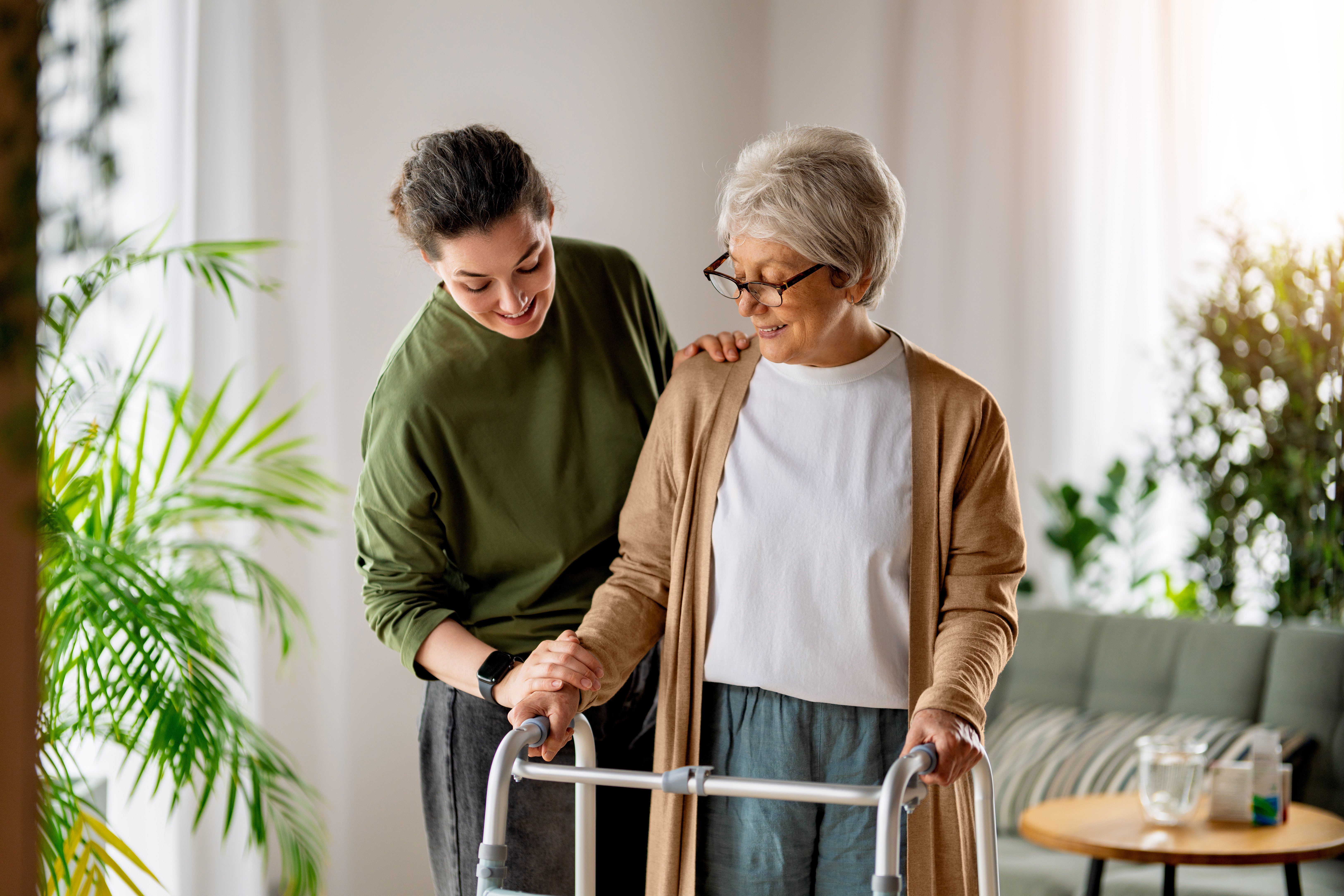 Heartwarming scene of a compassionate young woman helping her elderly mother walk with a mobility aid inside a cozy, sunlit home. The supportive relationship showcases love, trust, and intergenerational family bonds, making this image ideal for concepts like home care, aging in place, healthcare support, and family values.